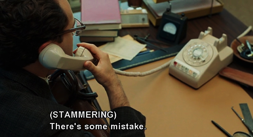 A frame from A Serious Man showing Larry Gopnik talking on the phone at his office, with the manner-of-speaking caption (STAMMERING).