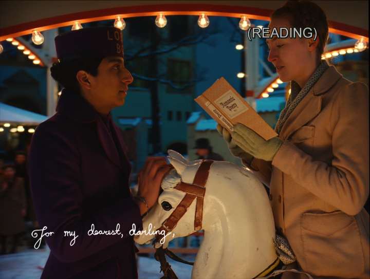 A frame from The Grand Budapest Hotel showing Zero and Agatha on a lighted carousel, with the closed caption (READING) stamped on Agatha's forehead.