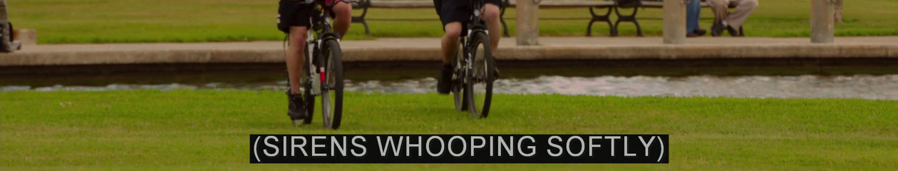 In this frame from 21 Jump Street (2012), the bottom half of two bicycle riders head towards the camera on park grass. The caption: (SIRENS WHOOPING SOFTLY)