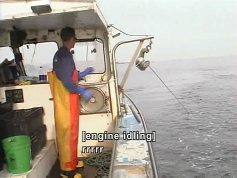 A fisherman in chest waders stands on the side of a small commercial fishing boat. The captions: [engine idling] and rrrrr