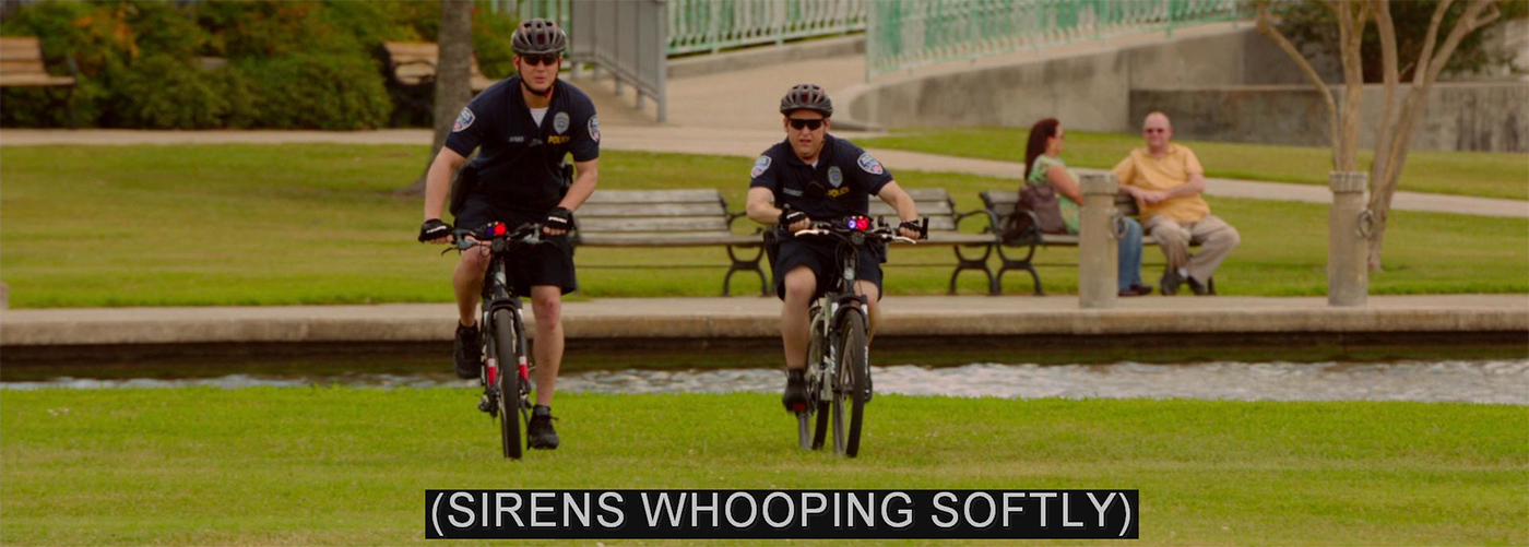 A frame from 21 Jump Street featuring Jenko and Schmidit riding bicycles on park grass. A small red light is visible on each bike's handlebars. The caption: (SIRENS WHOOPING SOFTLY)